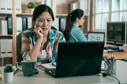 young concentrated asian woman employee indoors working with laptop computer thinking frowning confused. Coworking concept. female designer staring at notebook pc screen considering new project.