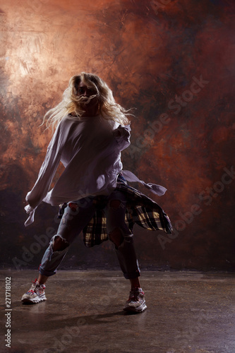 Photo of young blonde dancer in torn jeans and sneakers on dark brown background © Sergey