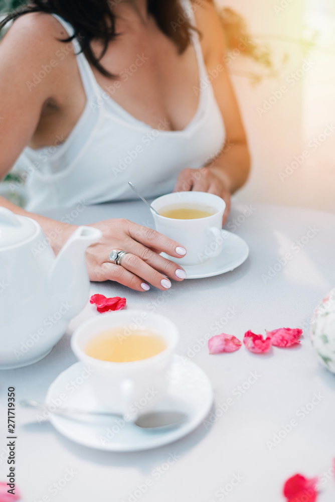Tea cups and woman's hands holding one cup on table with rose petals, top view. Tea cups and woman's hands holding one cup on table with rose petals, top view.