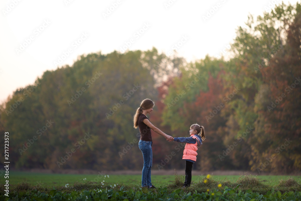 Fototapeta premium Back view of young slim attractive mother and child girl standing in green meadow holding hands outdoors on forest trees blurred background.