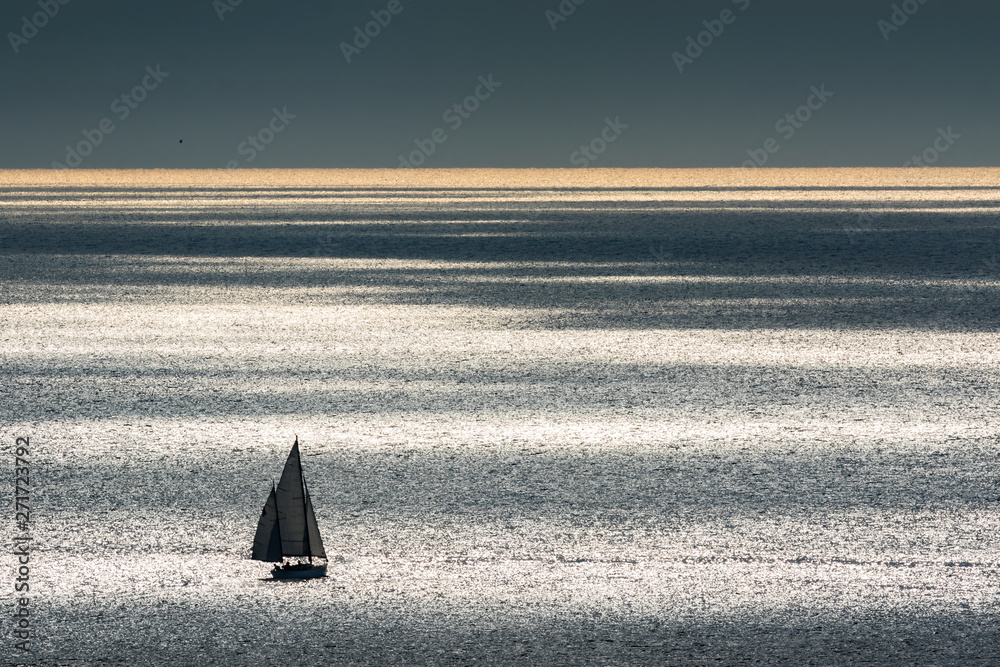 A sailing ship with two masts sailing off the coast in Spain in the Mediterranean Sea. The sun in the backlight forms glittering water with many different colors. The ship forms a silhouette. Aerial v