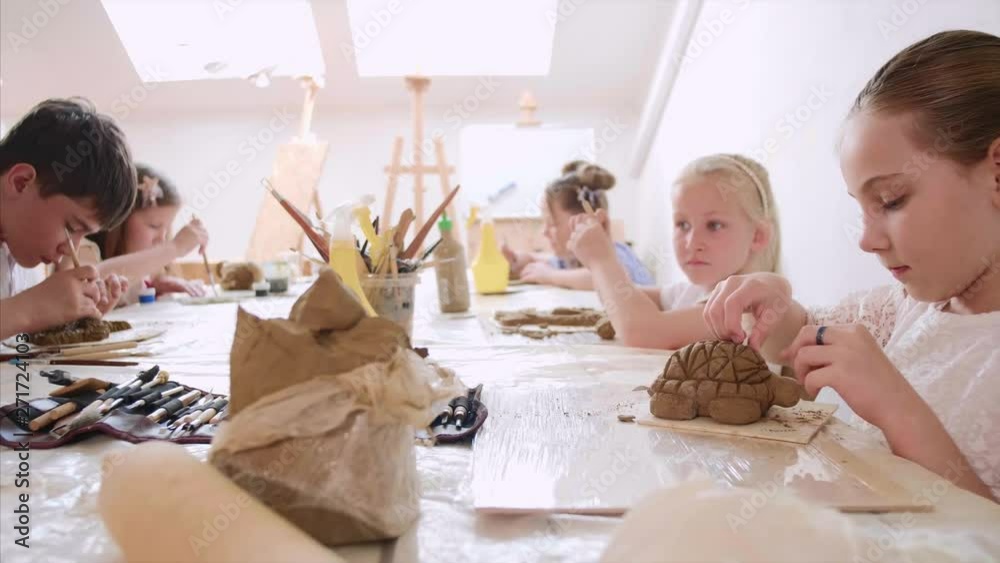 Children are sculpting toys from wet clay. They are using special tools under the guidance of a teacher on art lesson in workshop in elementary school.