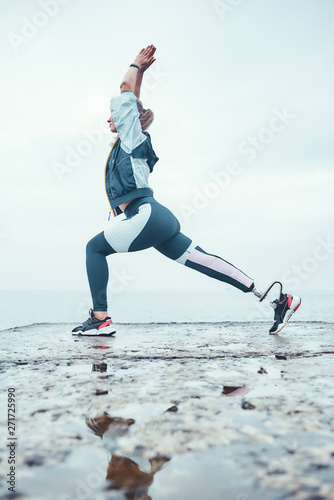 Surya Namaskar. Vertical photo of disabled athlete woman in sportswear with prosthetic leg standing in yoga pose on the stone in front of the sea.