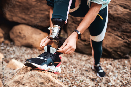 Cropped image of disabled athlete young woman in sports clothing with prosthetic leg tying shoelace while standing at the beach photo