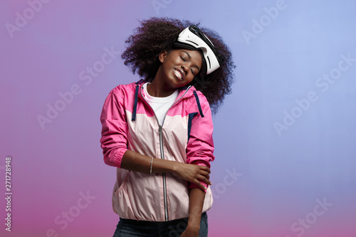 Funny curly brown-haired girl dressed in the pink sports jacket is wearing on her head the virtual reality glasses in the studio on neon background photo