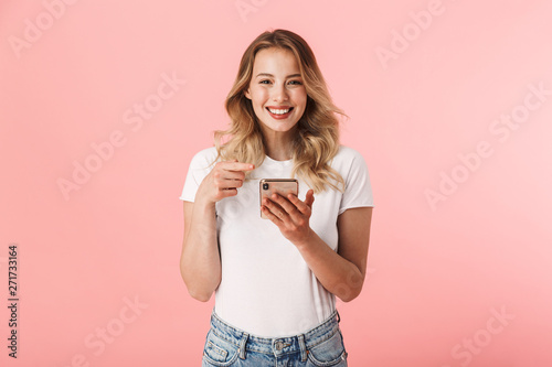 Happy young blonde woman posing isolated over pink wall background using mobile phone.