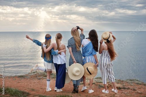 Five attractive and fun young girls having fun on the beach. Teenage sisters are resting together on the rocky shore of the blue ocean on a cloudy day against the blue sky and the coastline.