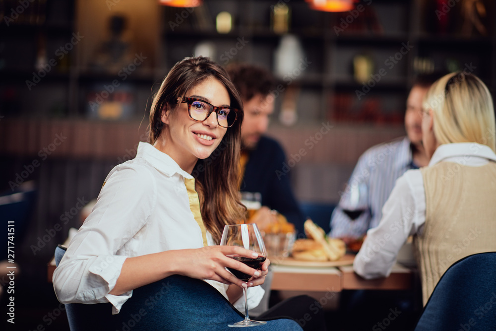 Portrait of gorgeous brunette looking at camera and holding glass with red wine while sitting at restaurat. In background friends eating diner.