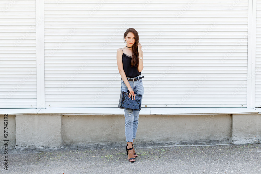 Young stylish woman wearing black cami silk top, blue cropped denim jeans, black high heel sandals and holding black handbag posing against white street wall. Trendy casual outfit. Street fashion.