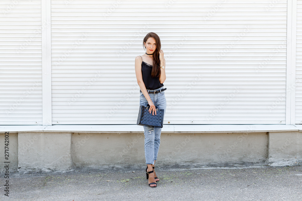 Young stylish woman wearing black cami silk top, blue cropped denim jeans, black high heel sandals and holding black handbag posing against white street wall. Trendy casual outfit. Street fashion. Young stylish woman wearing black cami silk top, blue cropped denim jeans, black high heel sandals and holding black handbag posing against white street wall. Trendy casual outfit. Street fashion.