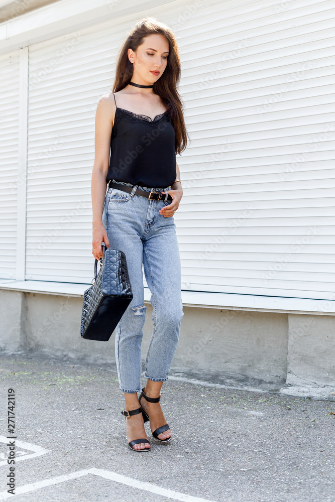 Young stylish woman wearing black cami silk top, blue cropped denim jeans,  black high heel sandals and holding black handbag walking against white  street wall. Trendy casual outfit. Street fashion. Stock Photo