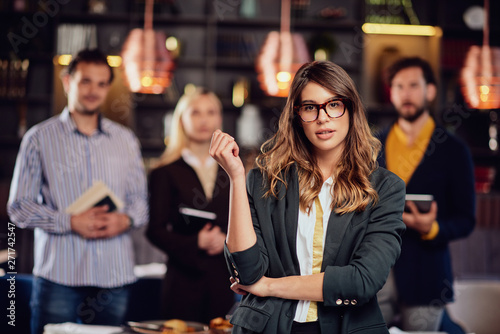 Serious Caucasian businesswoman with long brown hait and dressed smart casual standing in restaurant. In background her successful team posing.