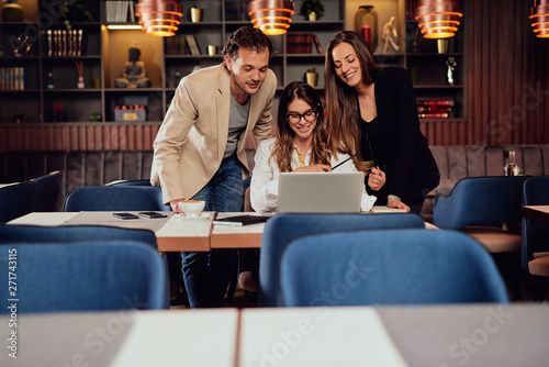 Charming buisnesswoman with brown hair and dressed elegant sitting at restaurant and looking at laptop. Next to her standing two colleagues and looking at laptop, too.