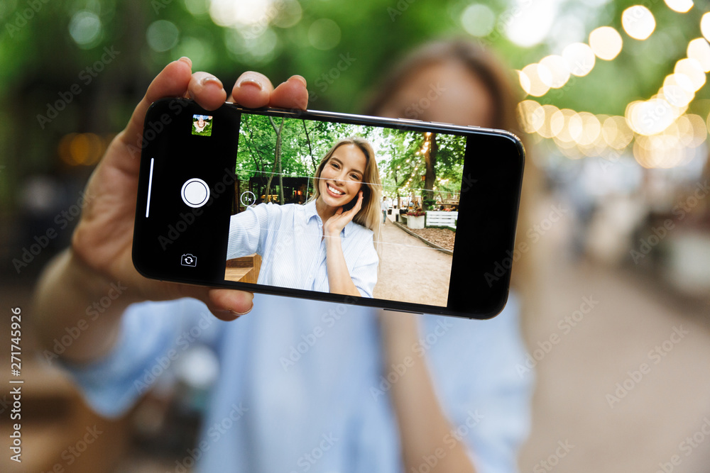 Excited happy young woman posing outdoors in park take a selfie by mobile phone.