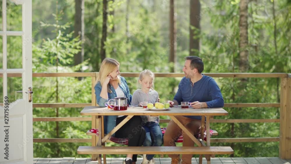 Happy family of mother, father and little daughter talking cheerfully while having breakfast at wooden table on country house porch surrounded by green trees in sunny morning, tracking right shot