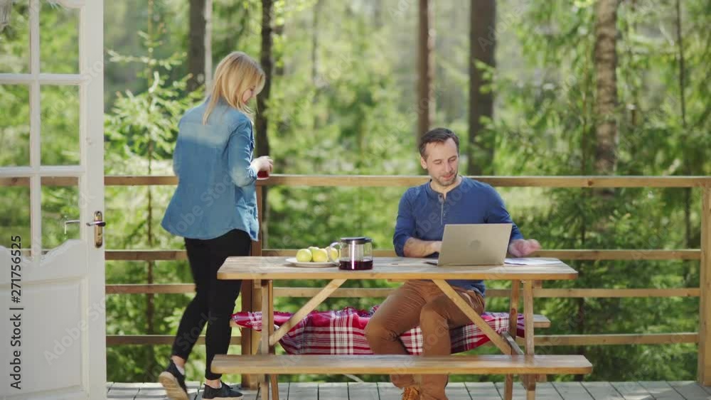 Concentrated businessman working on laptop computer on country house porch. Beautiful wife bringing tea for him. Cheerful couple talking at wooden table outdoors
