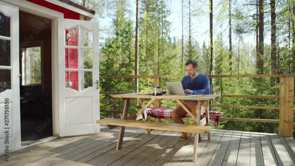 Tracking right shot of smiling middle aged man typing on laptop computer and drinking tea sitting at wooden table on country house porch surrounded by pine trees in summer Tracking right shot of smiling middle aged man typing on laptop computer and drinking tea sitting at wooden table on country house porch surrounded by pine trees in summer