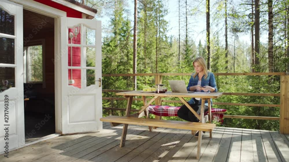 Tracking right shot of smiling young businesswoman working on laptop computer and making notes on paper sitting at wooden table on country house porch on summer day Tracking right shot of smiling young businesswoman working on laptop computer and making notes on paper sitting at wooden table on country house porch on summer day