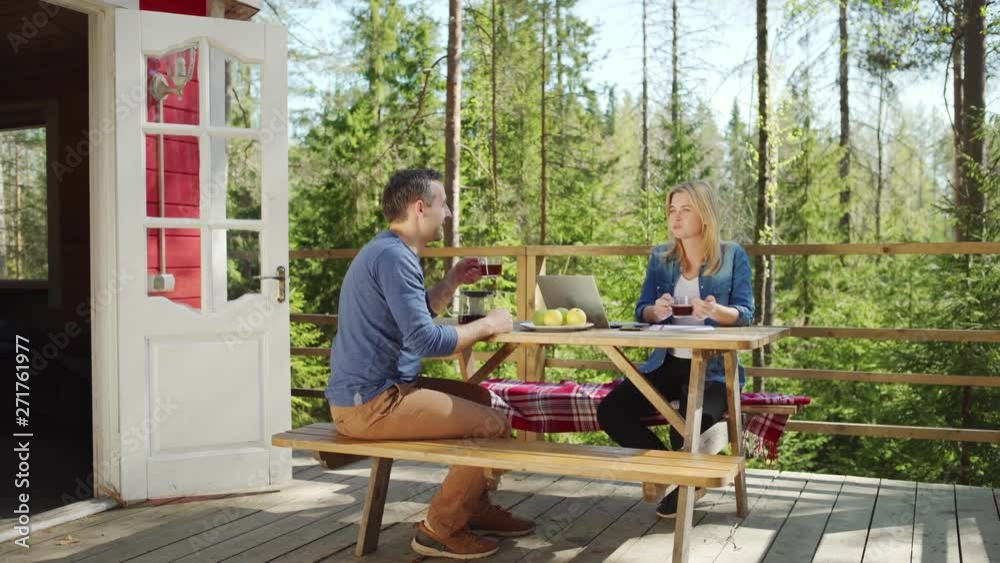 Tracking right shot of couple sitting at wooden table on country house balcony, drinking tea and talking. Open laptop, smartphone, papers and apples in front of them Tracking right shot of couple sitting at wooden table on country house balcony, drinking tea and talking. Open laptop, smartphone, papers and apples in front of them