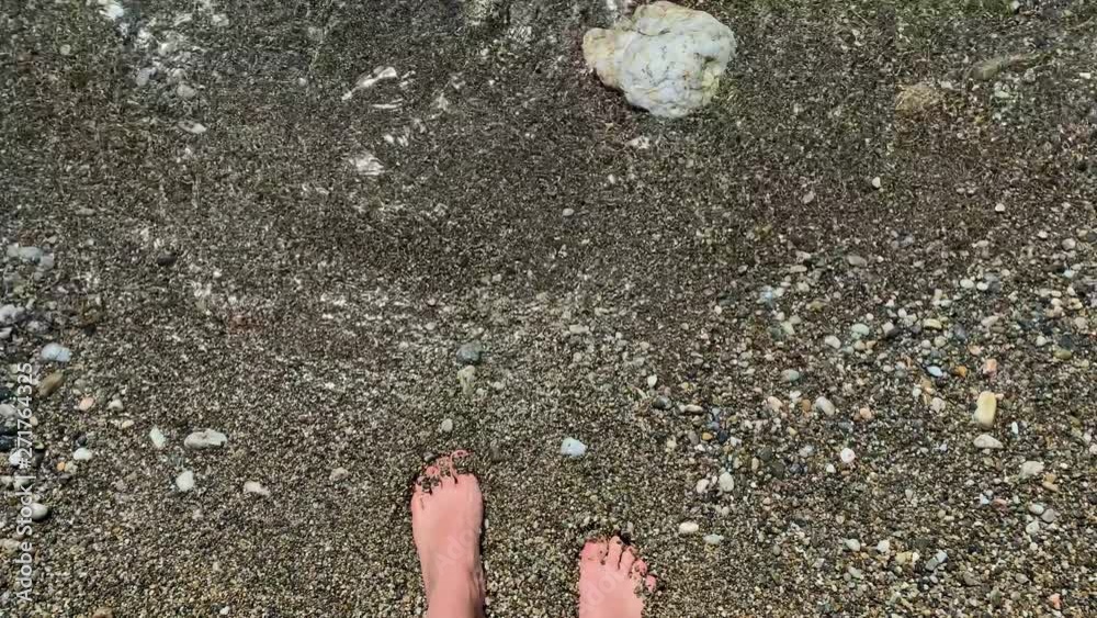Female feet standing on the pebble beach. Closeup of Sea waves washing over female feet. Point of view of beautiful young woman touching sea water. Girl on the stony beach on a sunny day. Female feet standing on the pebble beach. Closeup of Sea waves washing over female feet. Point of view of beautiful young woman touching sea water. Girl on the stony beach on a sunny day.