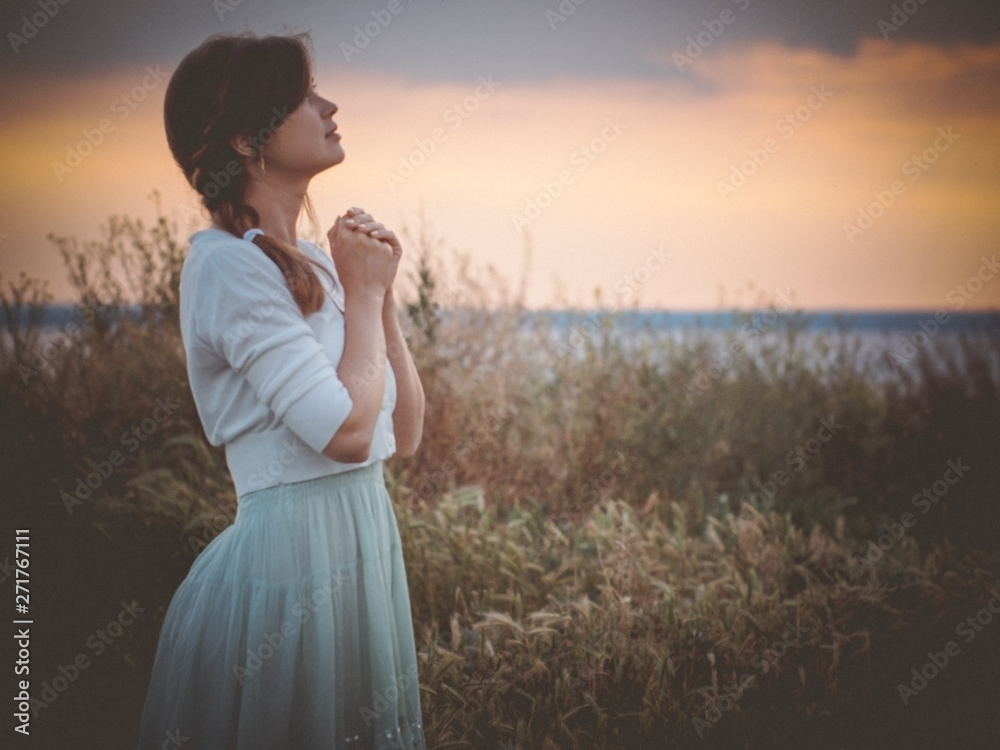 silhouette profile of a beautiful girl in a dress praying to God in the field, a young woman walking on nature during sunset looking up into the sky, religion concept