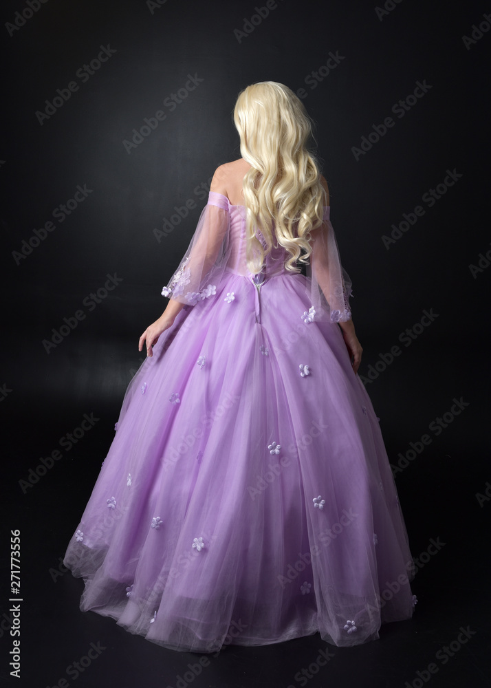 Fototapeta premium full length portrait of a blonde girl wearing a fantasy fairy inspired costume, long purple ball gown with fairy wings, standing pose on a dark studio background.