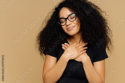 Touched lovely female with Afro hairstyle, keeps both palms on chest, has eyes shut from pleasure, hears heart piercing story, wears spectacles and casual t shirt, models against beige background photo