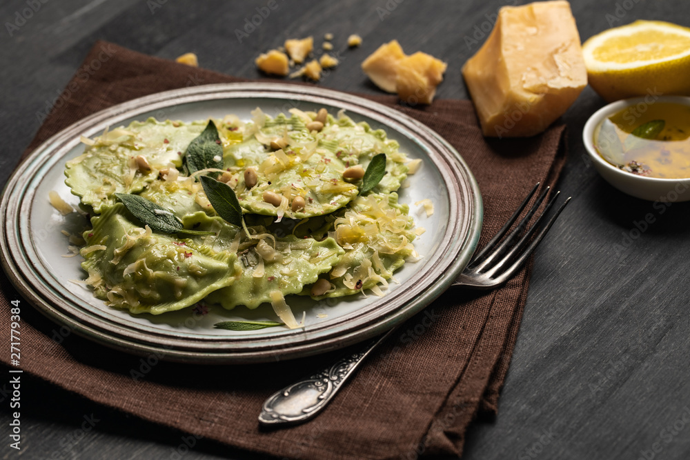 delicious green ravioli with sage, cheese and pine nuts served on black wooden table with fork, lemon and napkin