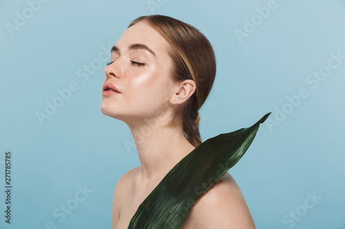 Beautiful young woman take care of her skin isolated over blue wall background posing with leaf flower.