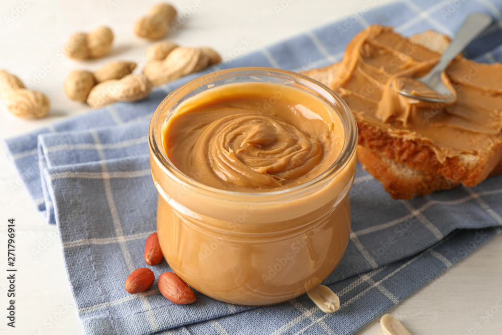 Glass jar with peanut butter, peanut, kitchen towel, spoon and peanut butter sandwich on white wooden background, space for text and closeup Glass jar with peanut butter, peanut, kitchen towel, spoon and peanut butter sandwich on white wooden background, space for text and closeup