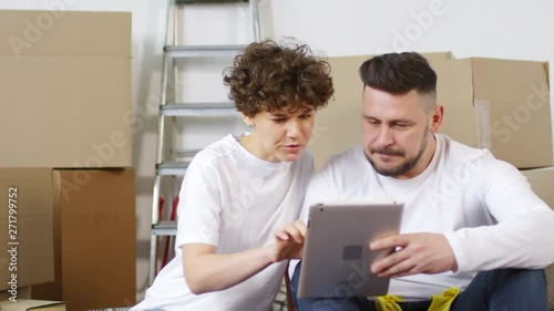 Panning thigh-up shot of middle-aged Caucasian couple sitting on floor in their new house among their unpacked possessions in cardboard boxes, browsing on tablet and planning interior redecoration
