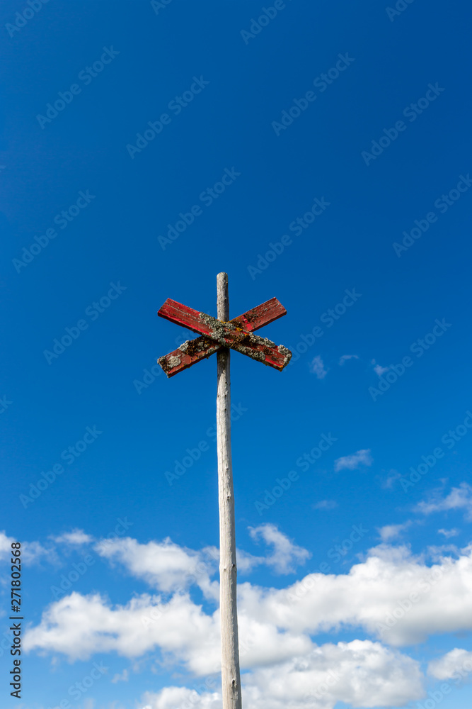 Old weathered wooden trail sign post with red cross against blue sky with clouds.