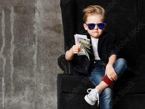 Portrait of self-confident rich kid boy siting in luxury armchair with bundles of dollars cash in his hand at text space photo