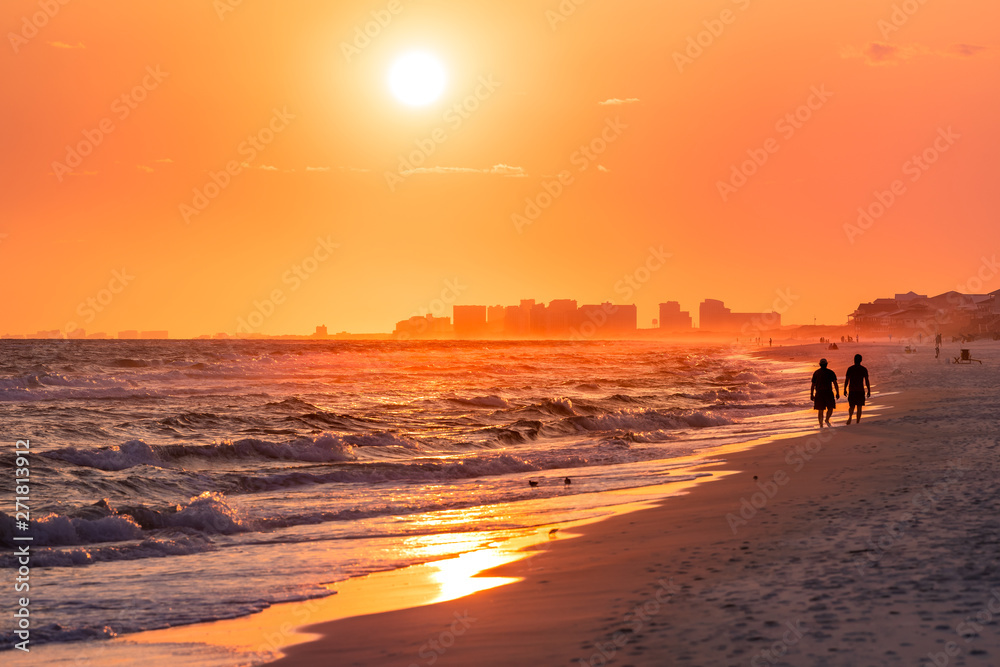 Couple walking on shore with dramatic orange red yellow sunset in Santa Rosa Beach, Florida with Pensacola coastline coast cityscape skyline in panhandle with ocean gulf mexico waves