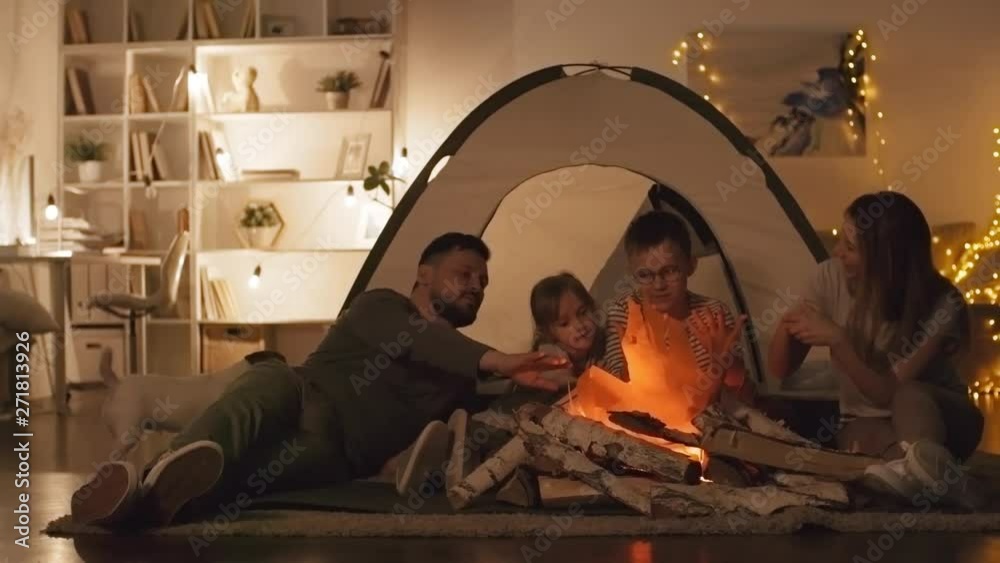Tracking right of Caucasian family couple and their two children sitting near artificial bonfire before tent in living room during nighttime while their dog going around