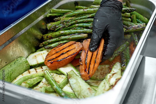 Grilled vegetables in a large container in the kitchen, the cook interferes with the hands of vegetables. Street market, street trading, healthy food. Selective focus. Close up photo