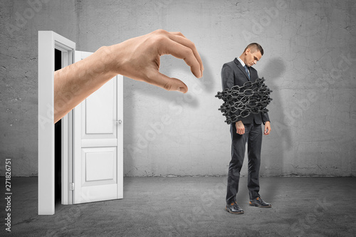 Full length side view of sad little businessman standing with chains around his body and huge hand emerging from door behind him. photo