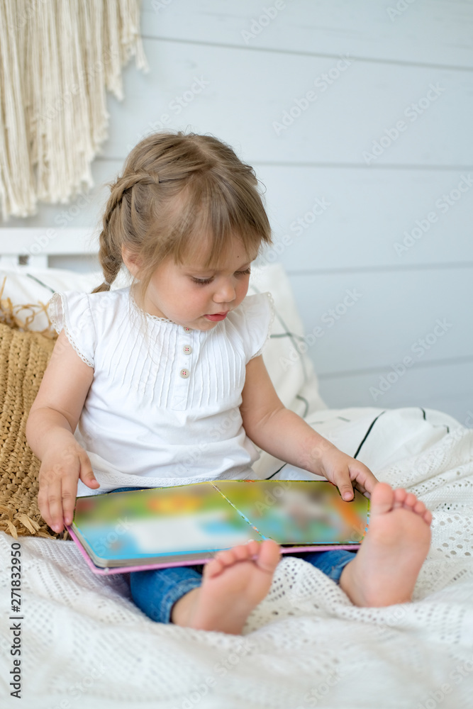 Beautiful little girl sitting on the bed and reading a book. The baby has blonde hair and blue eyes. White bedroom, cozy house. Teaching children to read.