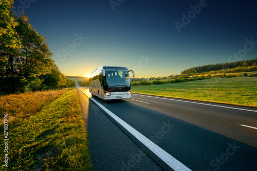 White bus traveling on the asphalt road around line of trees in rural landscape at sunset photo