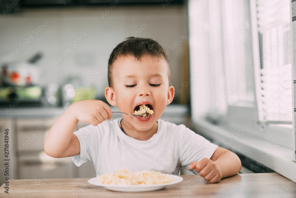 a child in the kitchen during the day eating pasta in a spiral in a white t-shirt a child in the kitchen during the day eating pasta in a spiral in a white t-shirt