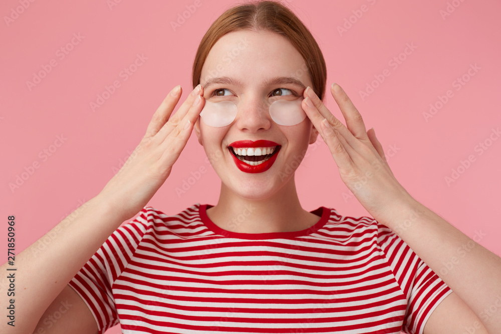 Cute red-haired lady in a red striped T-shirt, with red lips , touches his face with fingers, expects magical action of patches from dark circles under his eyes, enjoys free time for self-care. Cute red-haired lady in a red striped T-shirt, with red lips , touches his face with fingers, expects magical action of patches from dark circles under his eyes, enjoys free time for self-care.