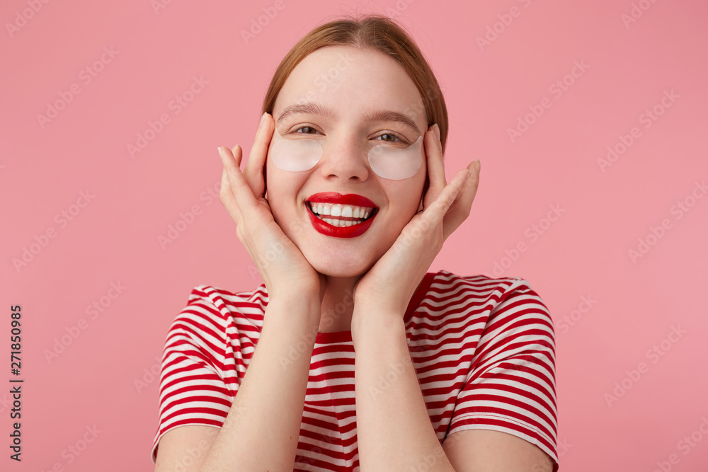 Nice young red-haired lady in a red striped T-shirt, with red lips , touches his face with fingers, very pleased with my new patches from dark circles under my eyes. Stands over pink background. Nice young red-haired lady in a red striped T-shirt, with red lips , touches his face with fingers, very pleased with my new patches from dark circles under my eyes. Stands over pink background.