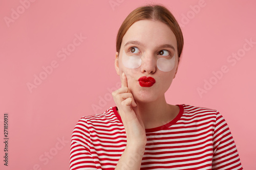 Nice red-haired young lady in a red striped T-shirt, with red lips , touches his cheek with finger, dreamily looks into the distance preparing for a date with his beloved. Stands over pink background.