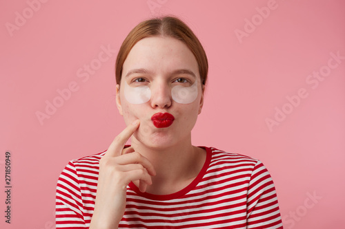 Portrait of thinking young red-haired girl with red lips and with patches under the eyes, wears in a red striped T-shirt, stands over pink background, points to his cheek and lokking at the camera.