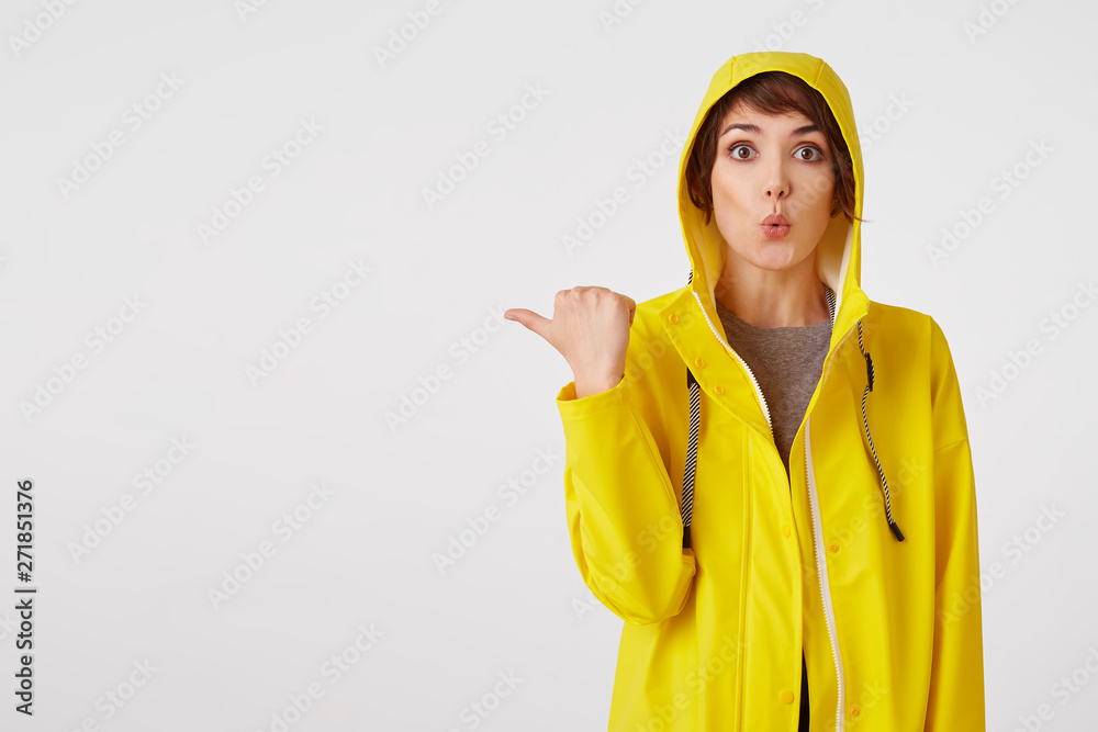 Young attractive girl in a yellow raincoat with a surprised expression on her face wants to draw your attention to the copy space on the left pointing with his finger, standing over white wall. Young attractive girl in a yellow raincoat with a surprised expression on her face wants to draw your attention to the copy space on the left pointing with his finger, standing over white wall.