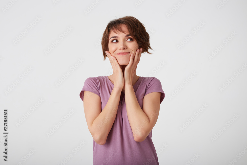 Young nice short-haired lady in blank t-shirt looks aside, touches cheeks, thinks about upcoming travels. Stands over white background. Young nice short-haired lady in blank t-shirt looks aside, touches cheeks, thinks about upcoming travels. Stands over white background.