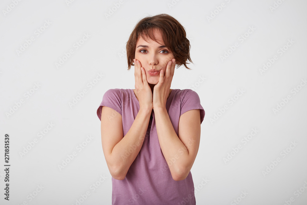 Young playful nice short-haired lady in blank t-shirt, looks at the camera and sedns kiss. Standing over white background. Young playful nice short-haired lady in blank t-shirt, looks at the camera and sedns kiss. Standing over white background.