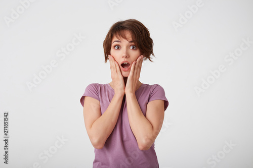 Portrait of wondered young attractive woman with a surprised expression on her face, standing over white wall with wide open moth and eyed. Positive emotion concept.