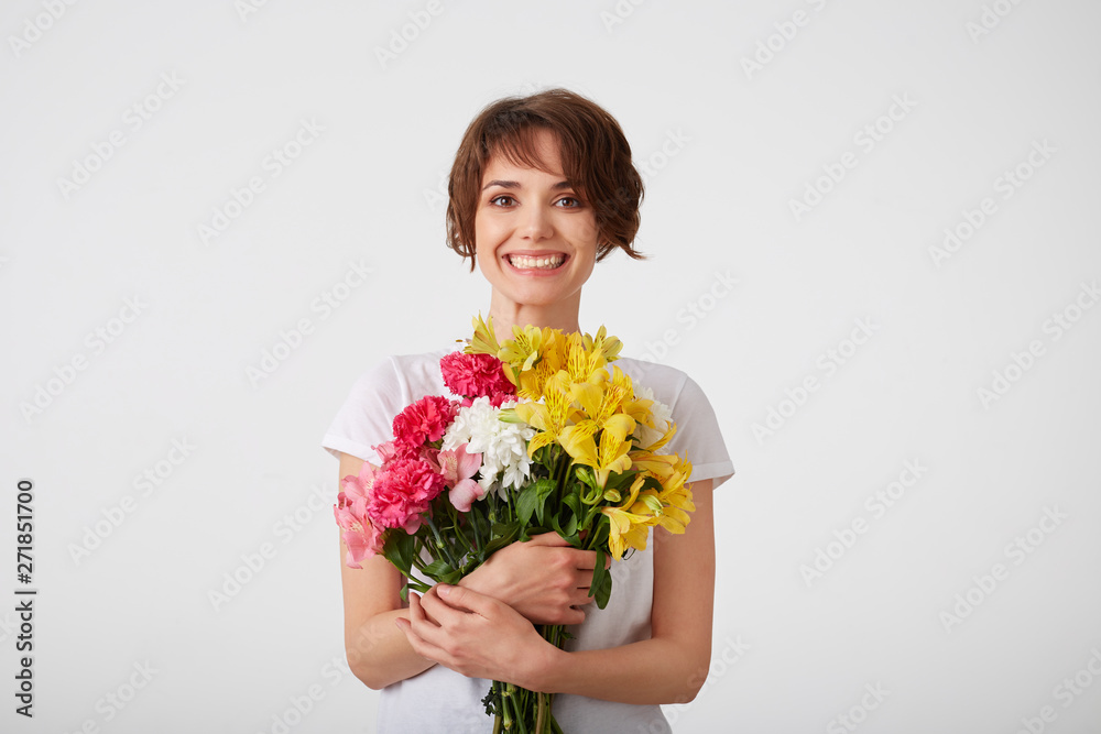 Portrait of happy smiling nice young short haired woman in white blank t-shirt, holding a bouquet of colorful flowers with closed eyes, standing over white background. Portrait of happy smiling nice young short haired woman in white blank t-shirt, holding a bouquet of colorful flowers with closed eyes, standing over white background.