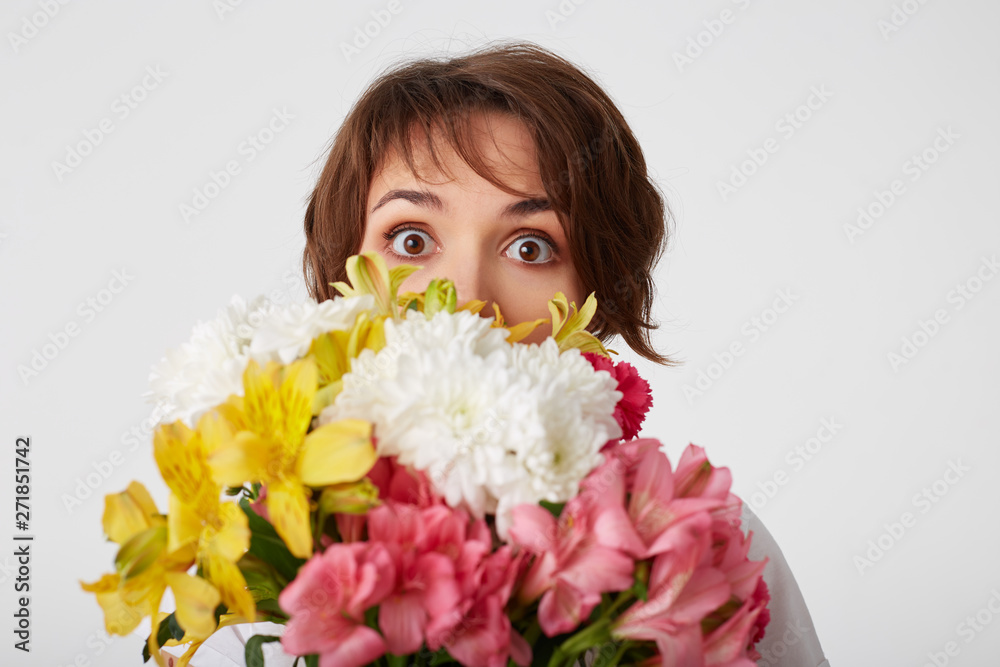 Portrait of nice short haired girl in white blank t-shirt, holding a bouquet, peeping from behind flowers, standing over white background with wide open eyes. Portrait of nice short haired girl in white blank t-shirt, holding a bouquet, peeping from behind flowers, standing over white background with wide open eyes.